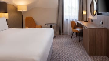 A hotel room with a large bed, a desk, two chairs, a small table, a TV, and a window with curtains.