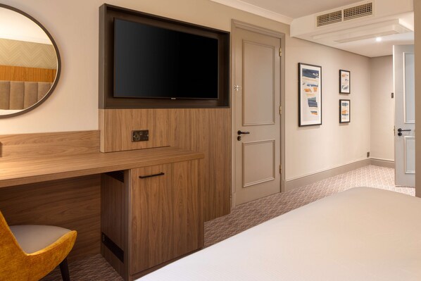 A hotel room with a bed, a desk with a chair, a television mounted on the wall, and framed pictures on the wall.