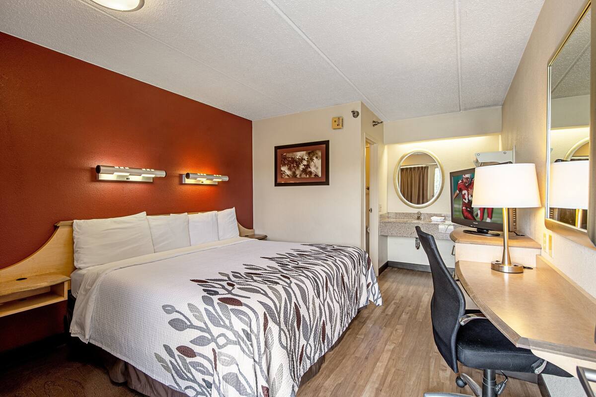 Una habitación de hotel con cama, escritorio, silla, televisor y espejo.