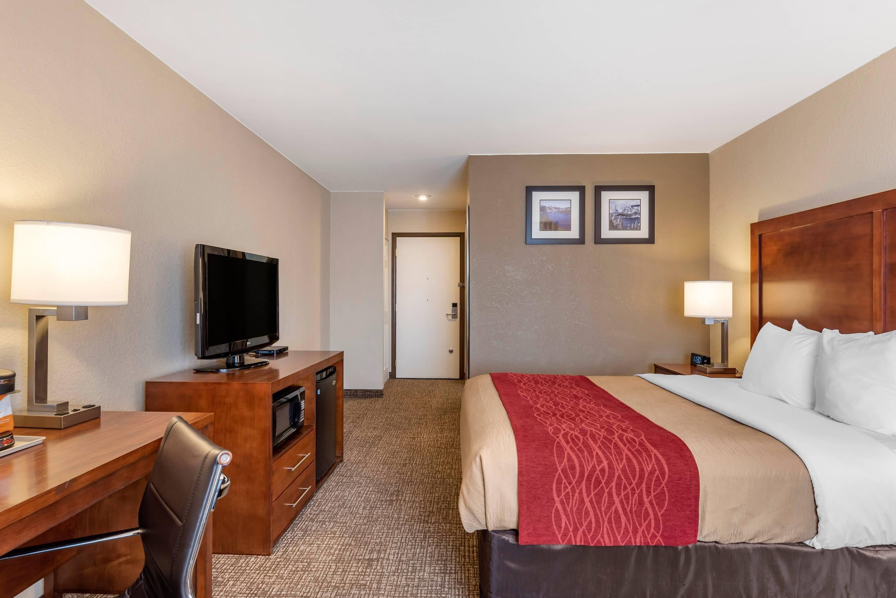 A hotel room with a bed, a desk with a television, a chair, a dresser, and two framed pictures on the wall.