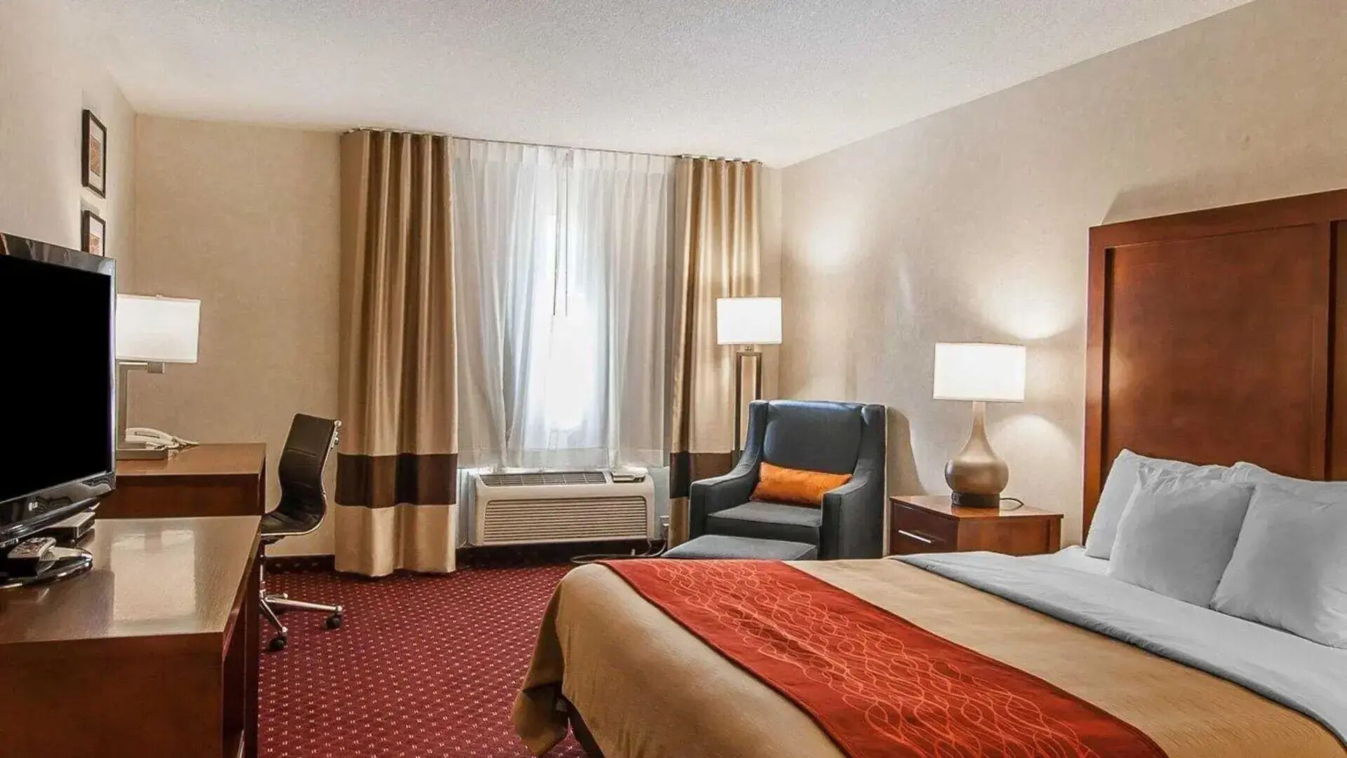 A hotel room with a bed, a desk with a TV, a chair, a lamp, and a window with curtains.