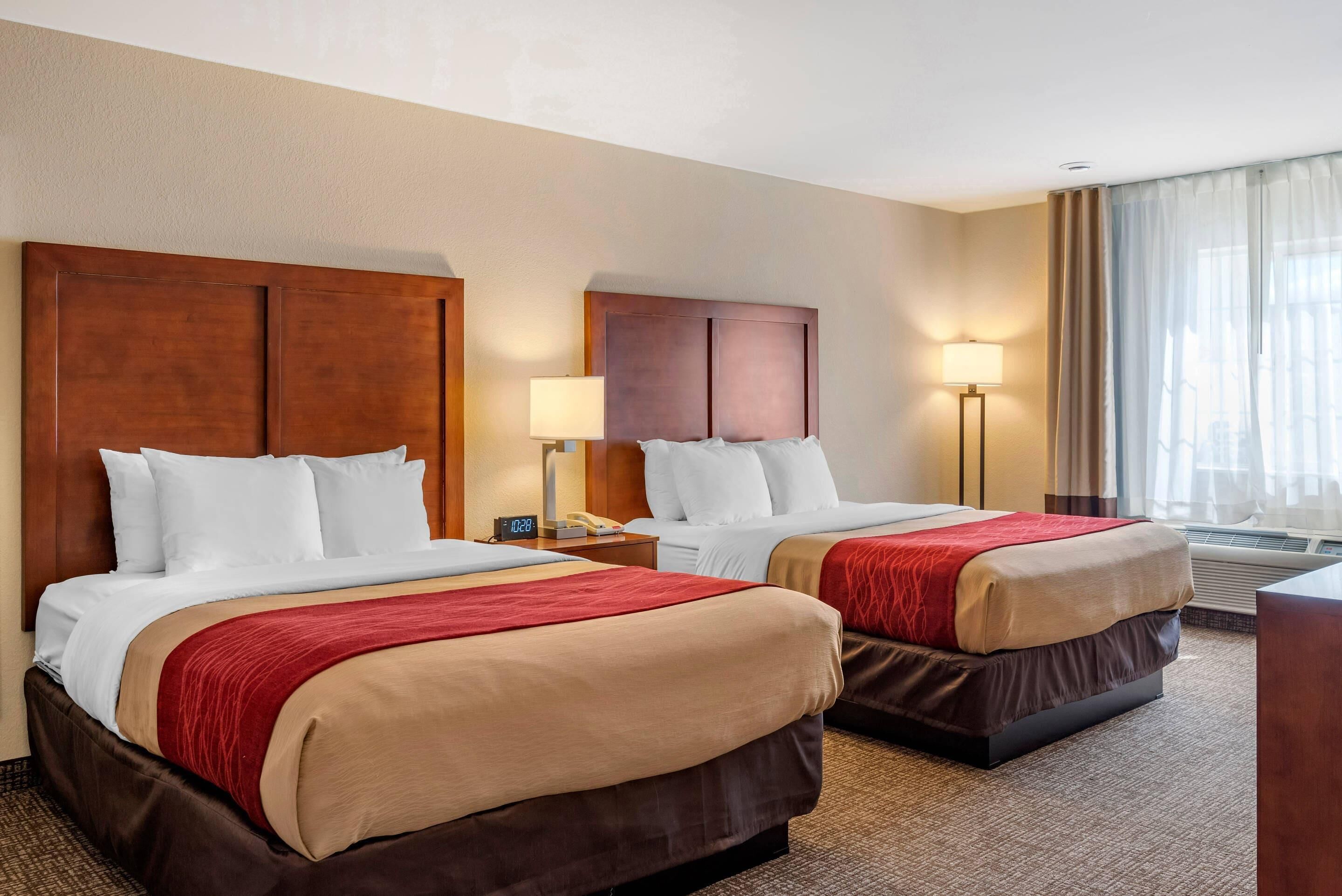 A hotel room with two beds, a nightstand with a lamp, a window with curtains, and an air conditioning unit.