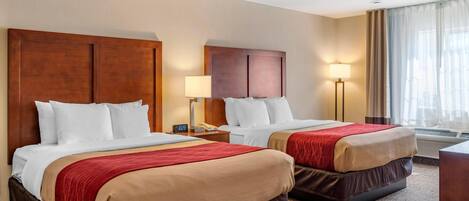 A hotel room with two beds, a nightstand with a lamp, a window with curtains, and an air conditioning unit.