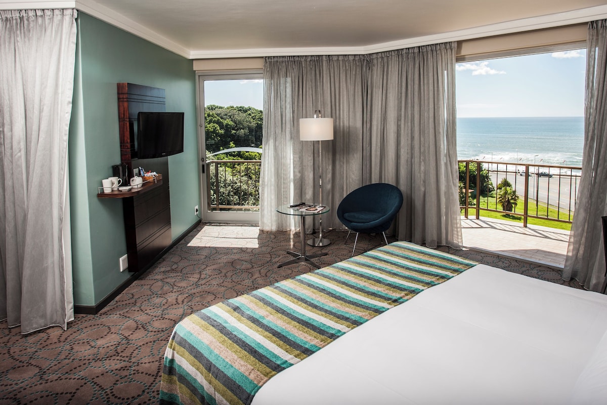 A hotel room with a bed, a TV, a chair, a table, and a balcony with a view of the ocean.