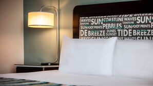 Una habitación de hotel con cama, mesita de noche, lámpara y una pared con una nube de palabras relacionadas con la costa.