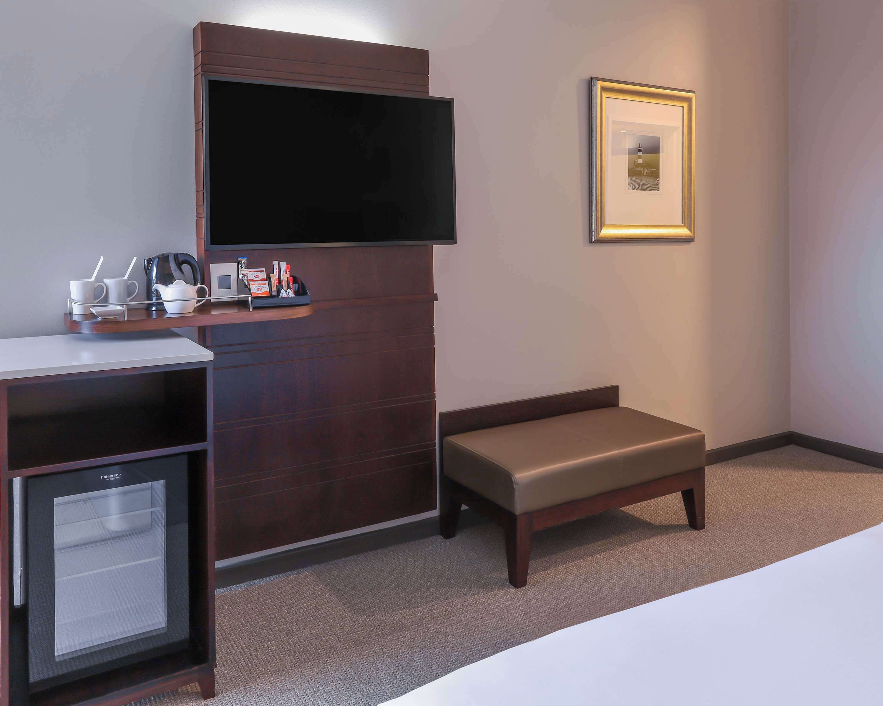 A hotel room with a flat-screen TV mounted on a wooden cabinet, a small bench, a desk with a coffee maker, and a framed picture on the wall.