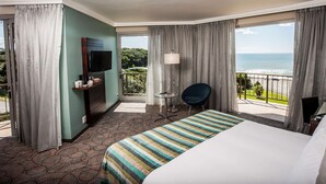 Habitación de hotel con cama, televisor, escritorio con silla y balcón con vista al océano.