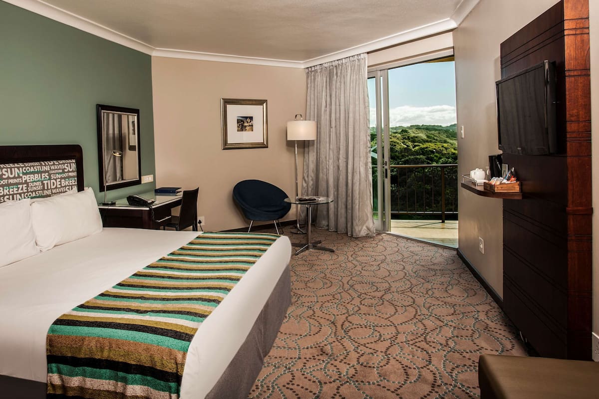 Une chambre d’hôtel avec un grand lit, un bureau, une chaise, une télévision et un balcon donnant sur un espace verdoyant.