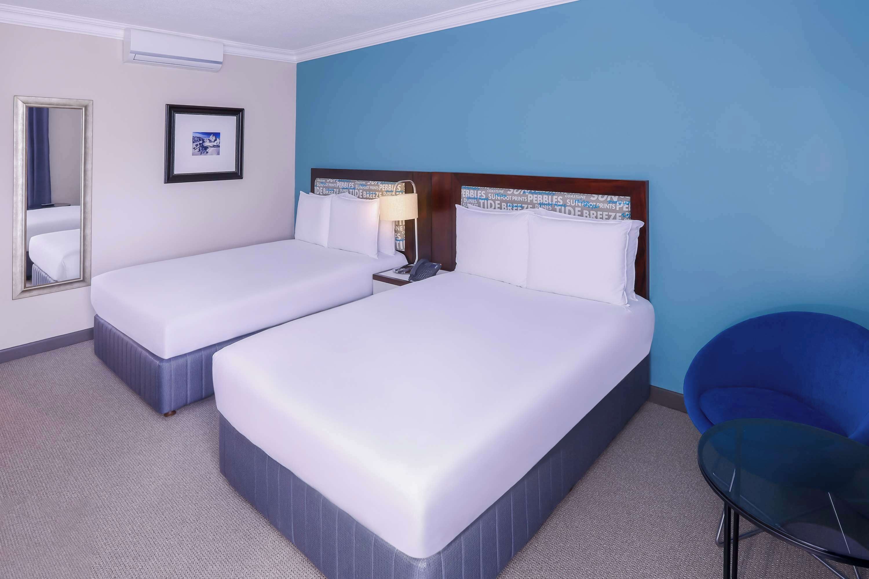 A hotel room with two beds, a bedside table with a lamp, a chair, and a mirror.
