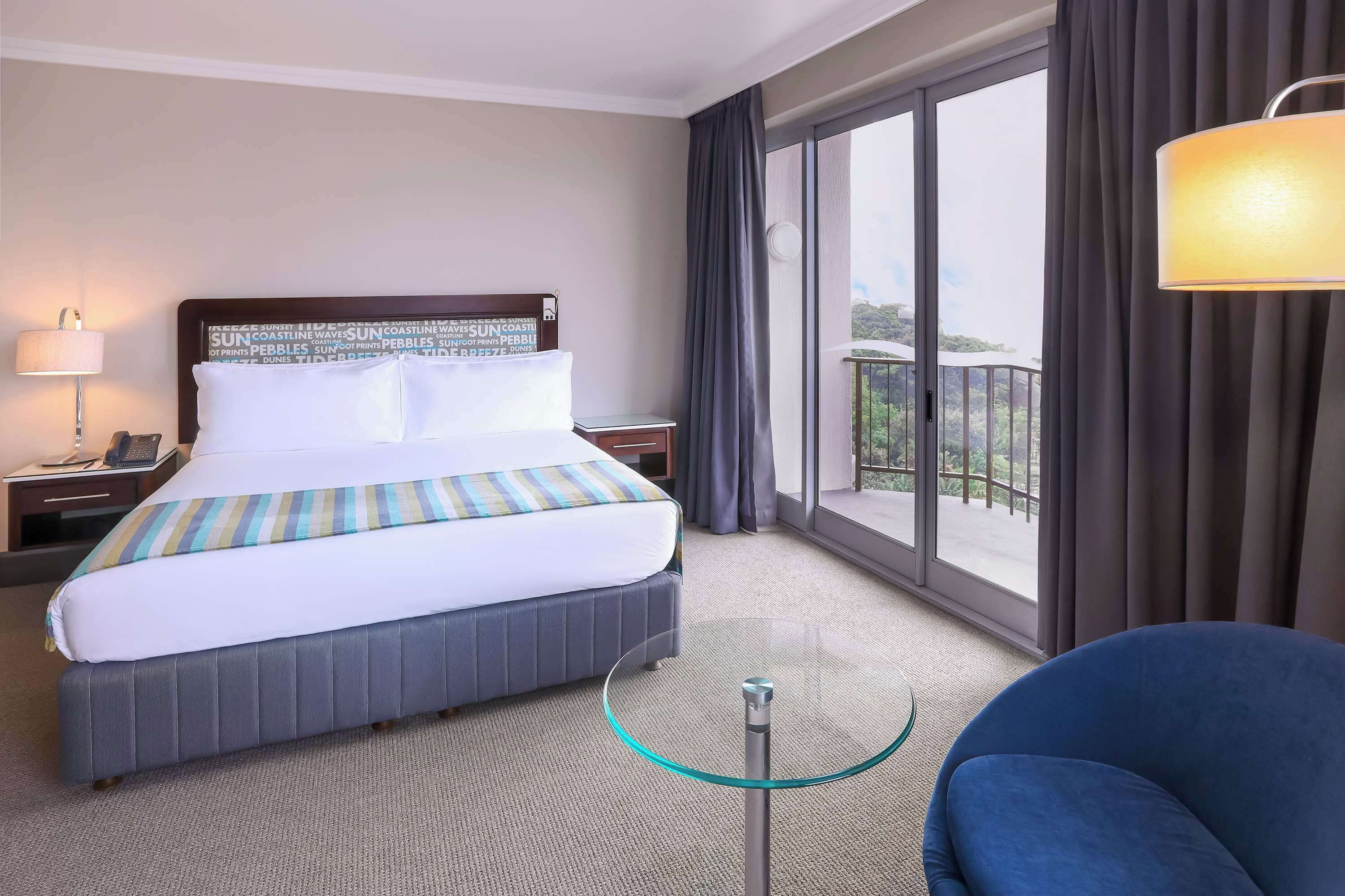 Habitación de hotel con una cama grande, una mesa pequeña, una silla azul y un balcón con cortinas.