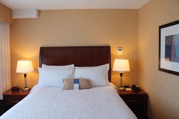 Una habitación de hotel con una cama grande, dos mesitas de noche, dos lámparas, una mesita auxiliar, un reloj y un cuadro en la pared.
