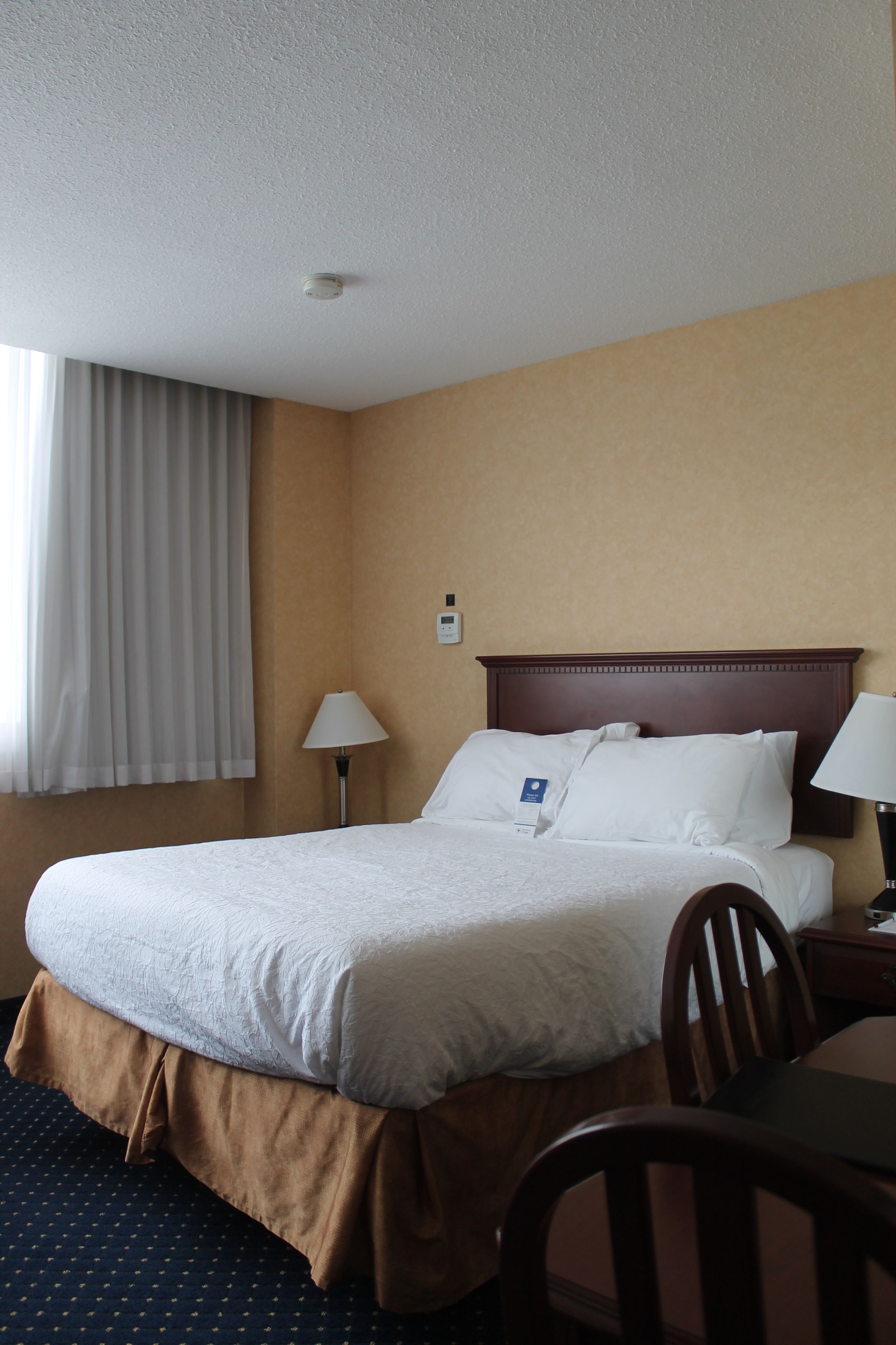 A hotel room with a large bed, a wooden headboard, a nightstand with a lamp, a chair, and a window with curtains.