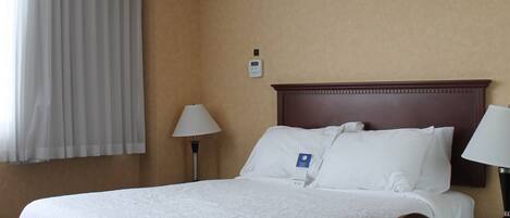 A hotel room with a large bed, a wooden headboard, a nightstand with a lamp, a chair, and a window with curtains.
