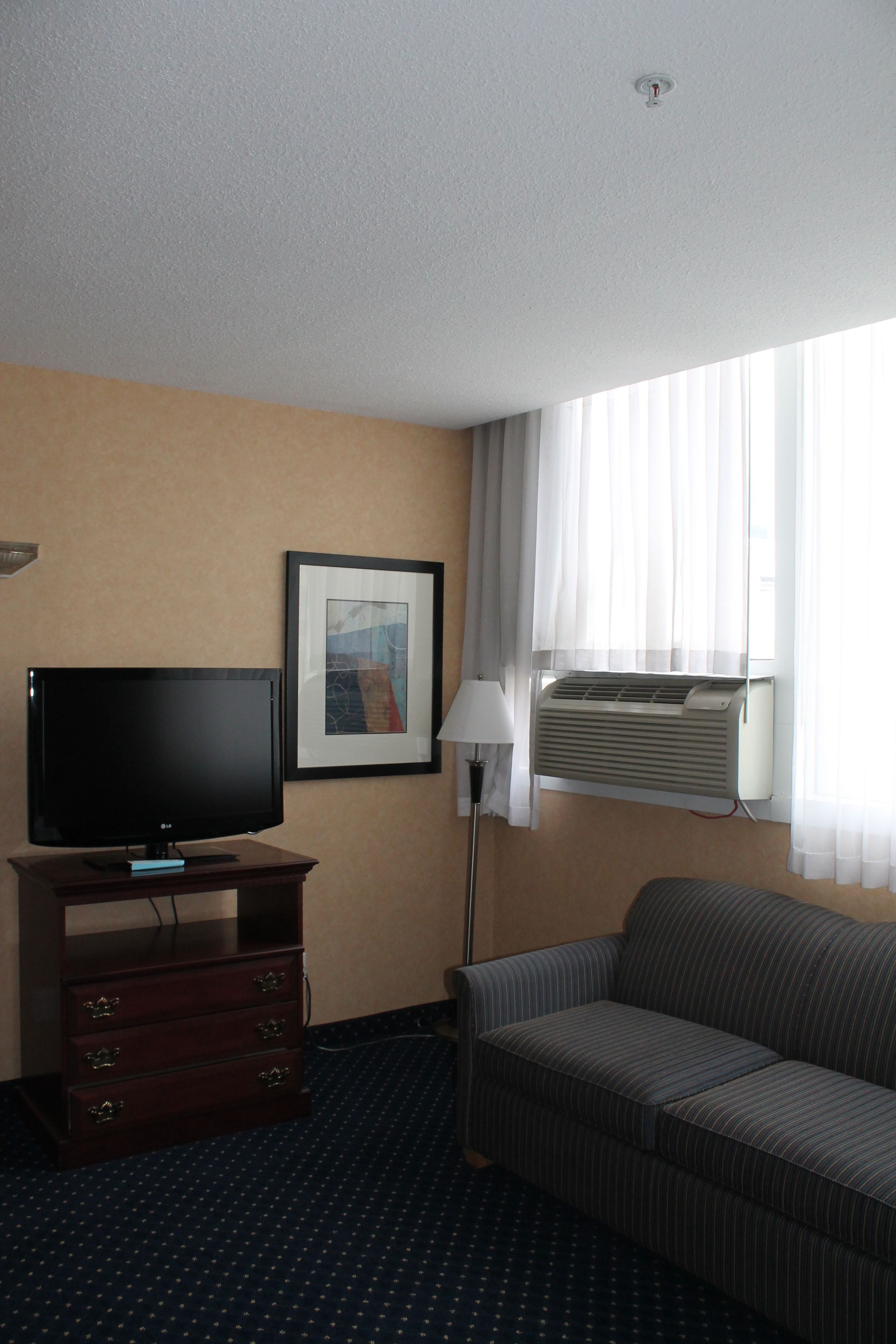 Habitación de hotel con televisor de pantalla plana sobre un soporte de madera, un cuadro enmarcado en la pared, una lámpara de pie, una ventana con unidad de aire acondicionado y un sofá a rayas.
