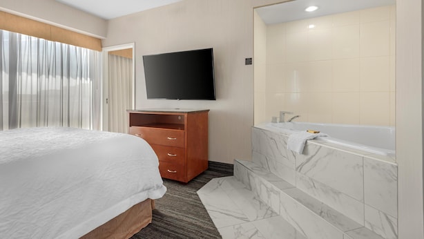 Habitación de hotel con cama, televisor montado en la pared, cómoda de madera y baño con bañera y baldosas de mármol.