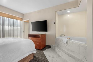 Habitación de hotel con cama, televisor montado en la pared, cómoda de madera y baño con bañera y baldosas de mármol.