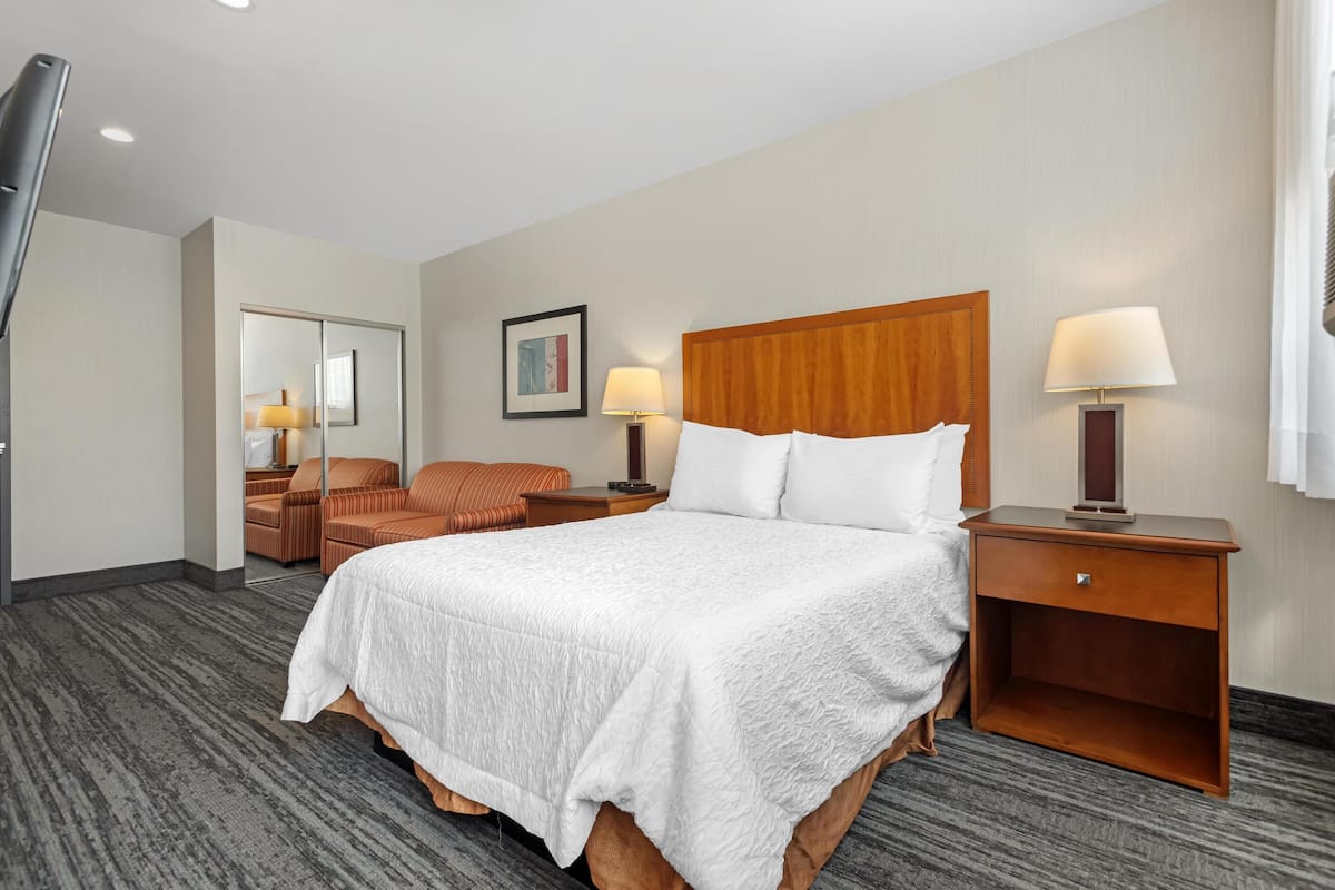 Habitación de hotel con una cama grande, dos mesitas de noche con lámparas, una silla, un espejo y un cuadro enmarcado en la pared.