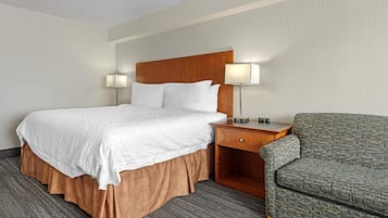 Quarto de hotel com uma cama grande, duas luminárias de cabeceira, uma mesinha de cabeceira de madeira e uma poltrona estampada.