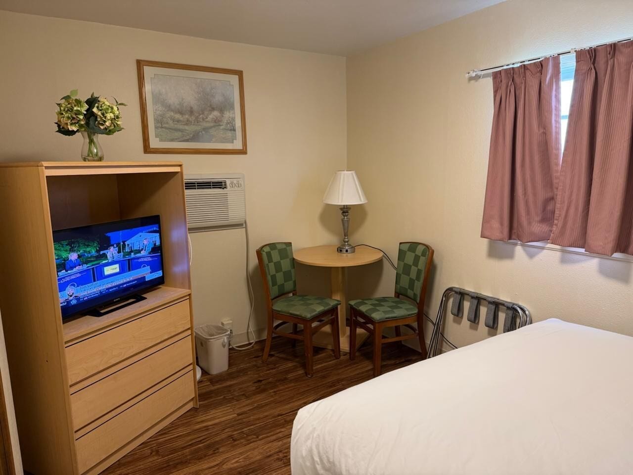 A hotel room with a bed, a TV on a wooden cabinet, a small table with two chairs, a lamp, and a window with curtains.