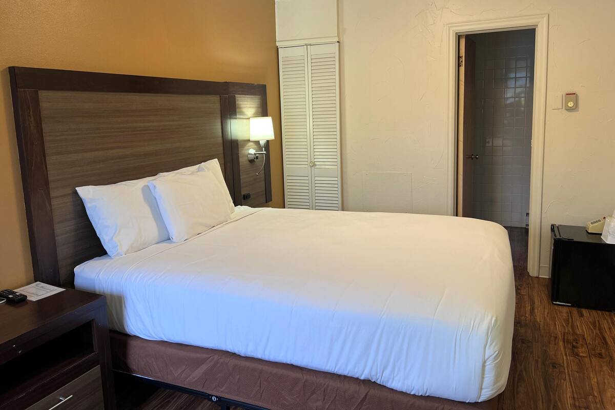 Una habitación de hotel con una cama grande, una mesita de noche con lámpara y una puerta que da a un baño.