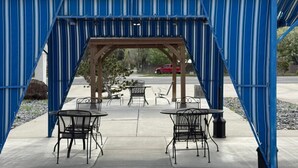 A covered outdoor seating area with tables and chairs, a wooden pergola structure, and a red car in the background. - Sunrise Motor Inn (Cody)