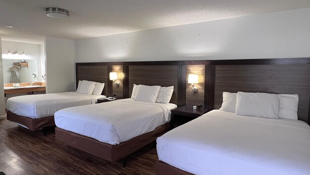 A hotel room with three beds, a wooden floor, a bathroom with a mirror and towel rack, and wall-mounted lights. - Sunrise Motor Inn (Cody)