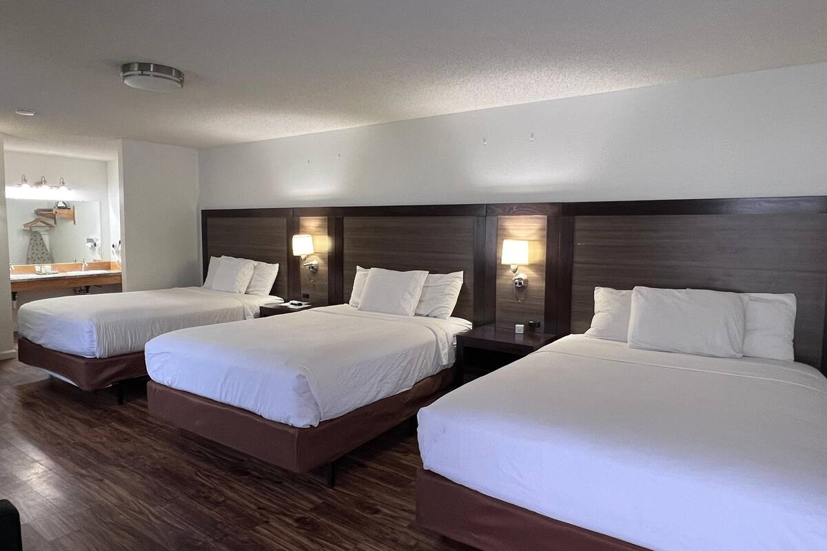Habitación de hotel con tres camas, suelo de madera, baño con espejo y perchero, y luces fijadas a la pared.