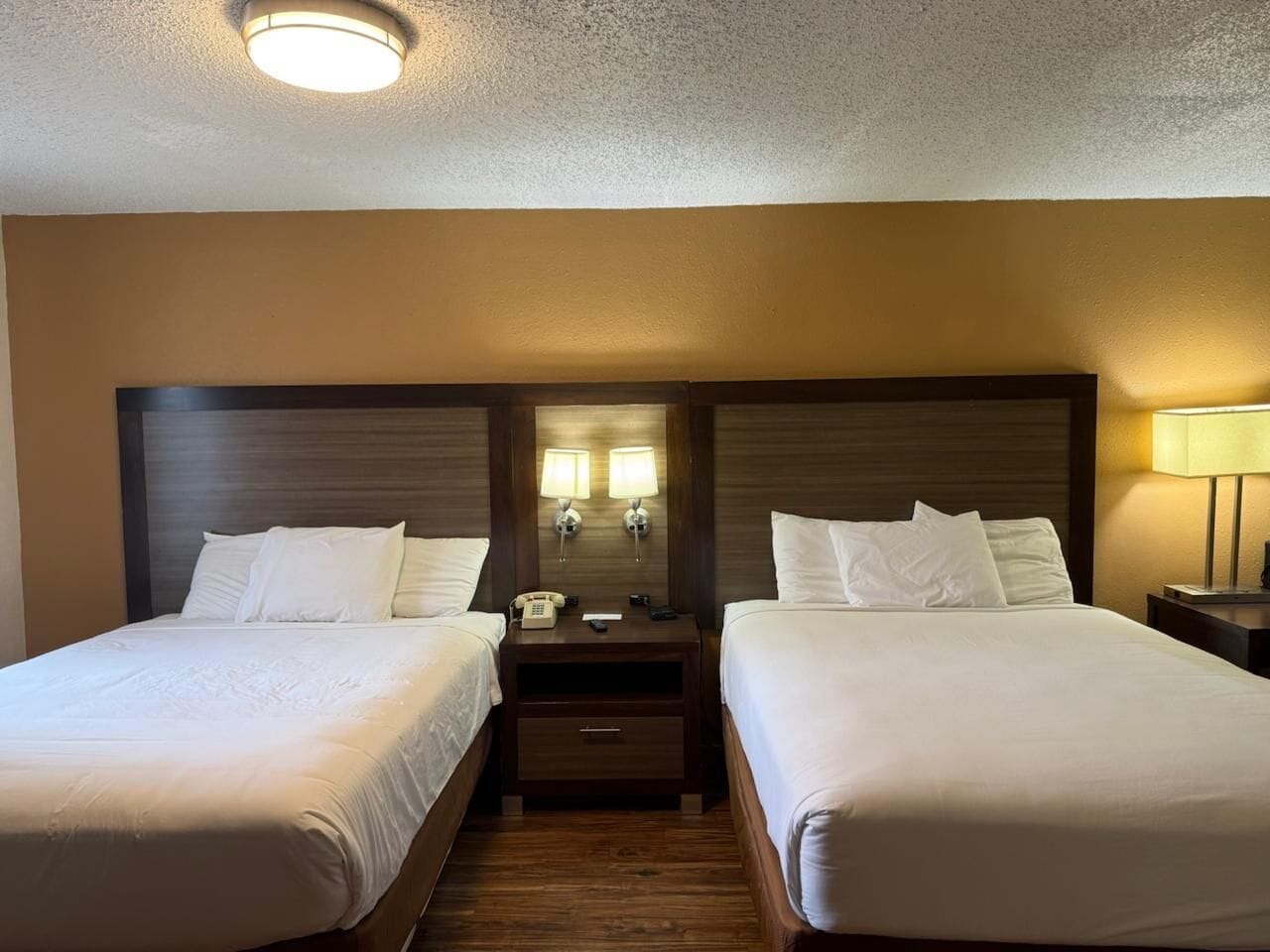 A hotel room with two single beds, a nightstand with a lamp, and a mirror above the nightstand.