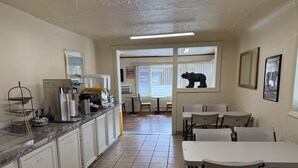 A kitchen with a coffee machine, a sink, and a window with a bear decoration. There are white chairs and a table in the dining area. - Sunrise Motor Inn (Cody)