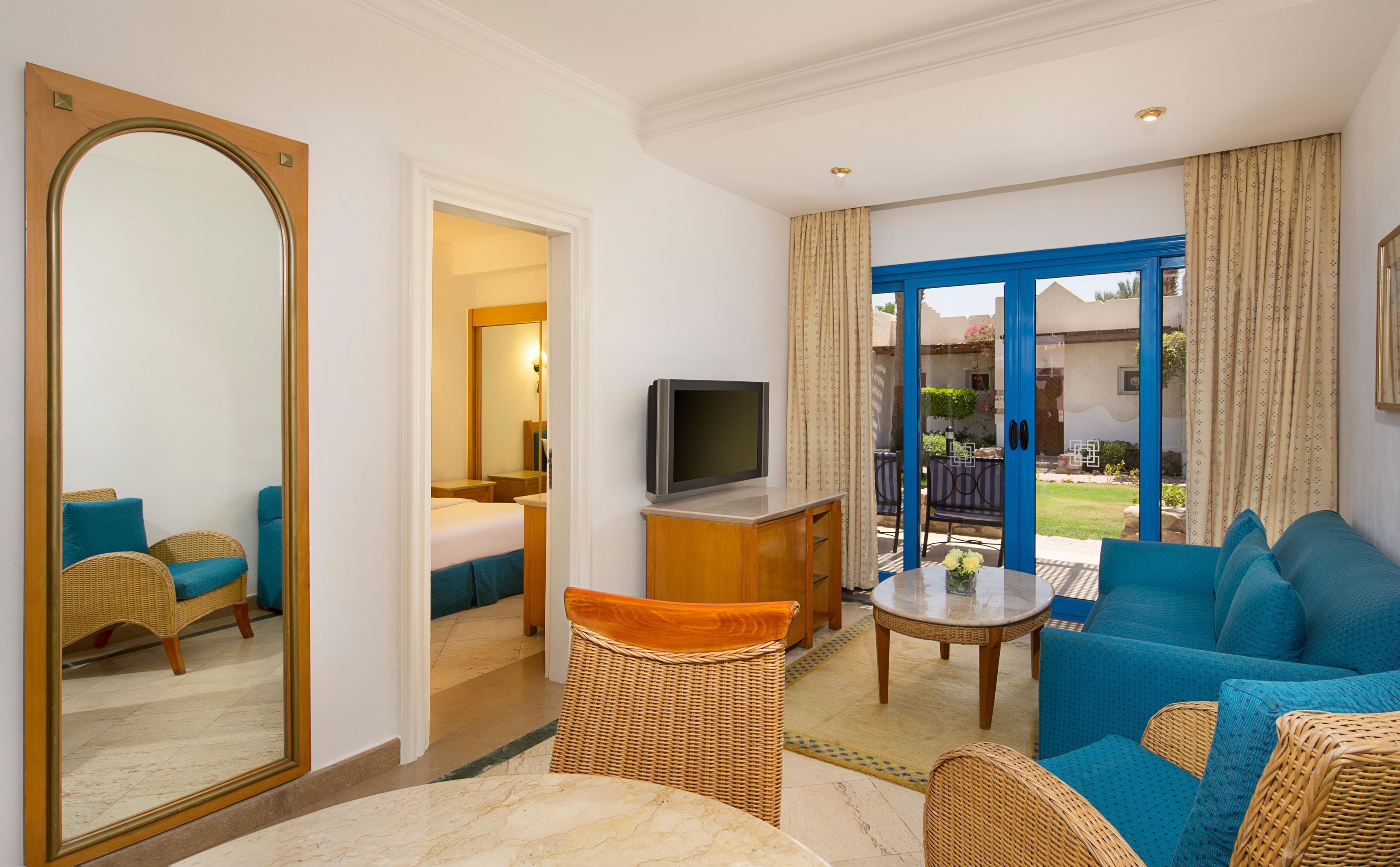 A hotel room with a bed, a TV, wicker furniture, a balcony with curtains, and a view of the outdoors.