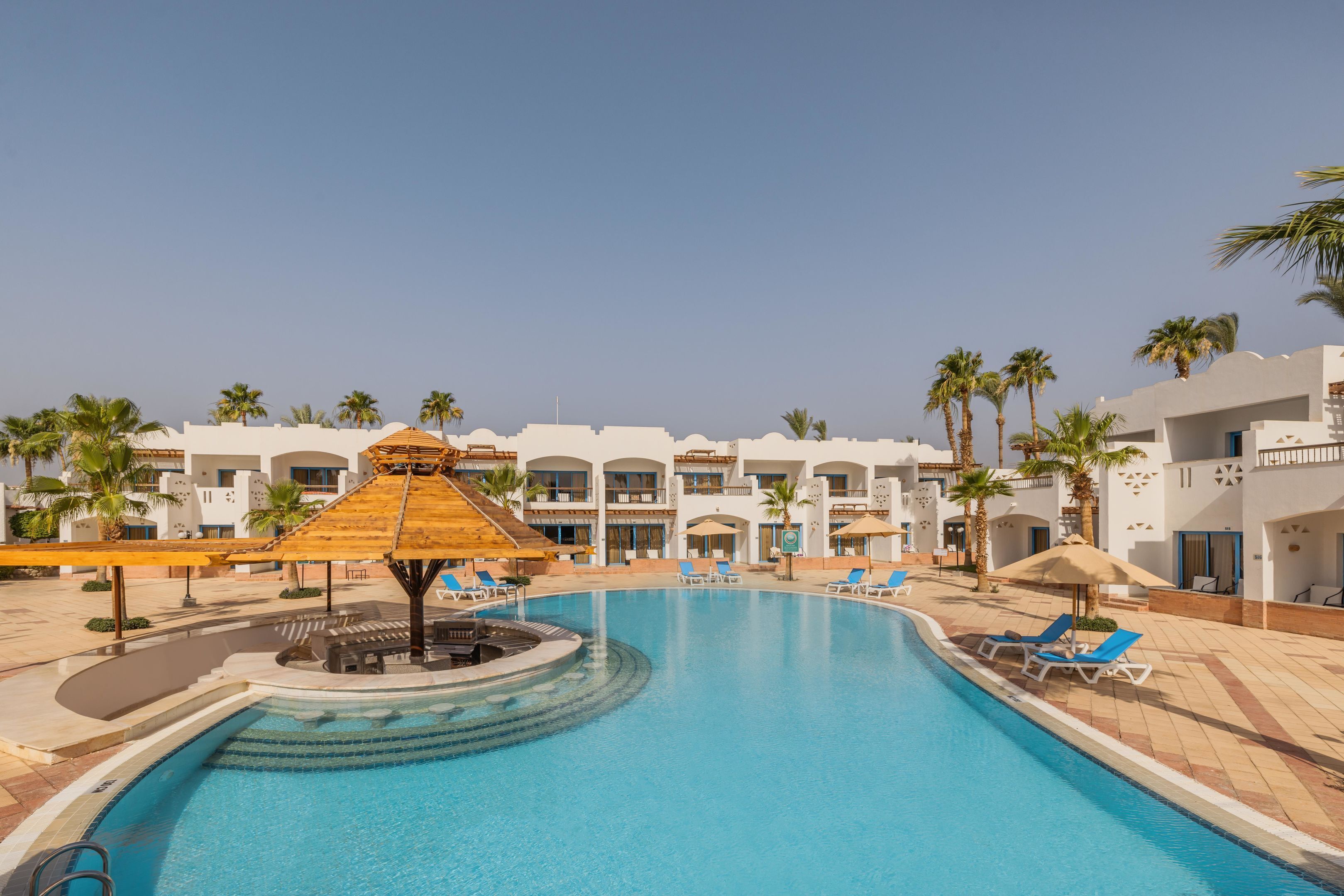 A swimming pool with lounge chairs and umbrellas, surrounded by palm trees and hotel buildings.