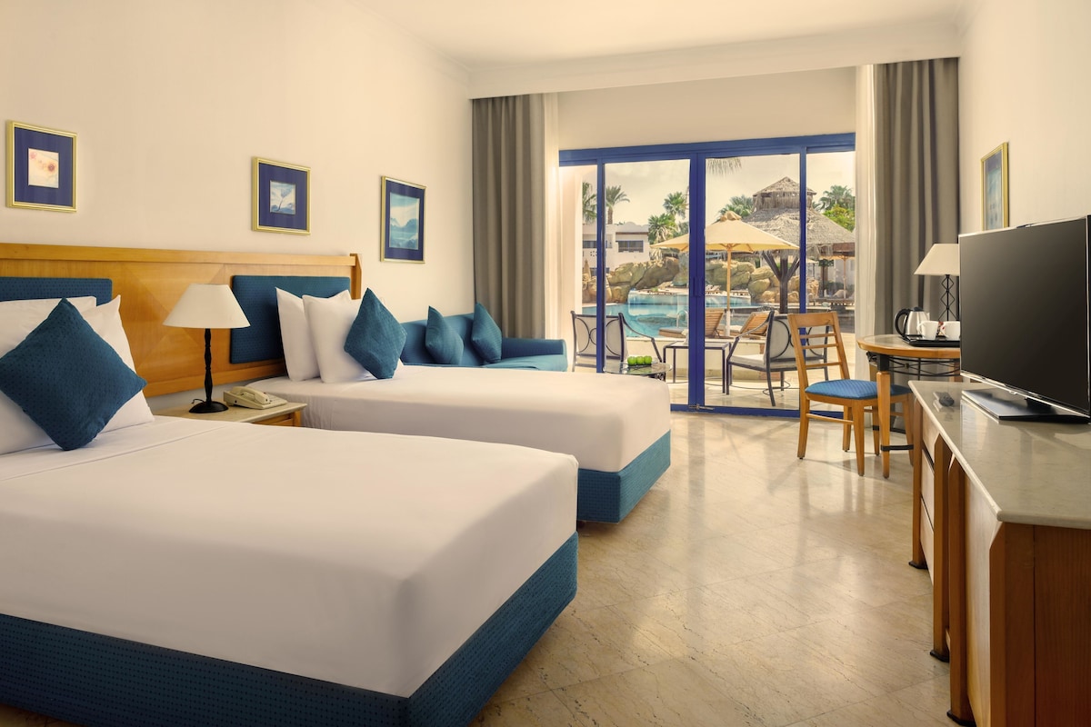 Une chambre d’hôtel avec deux lits, un bureau équipé d’une télévision, une chaise et une vue sur une piscine et un espace extérieur.