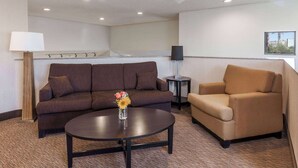 A hotel room with a brown sofa, a beige armchair, a round table with a vase of flowers, a standing lamp, and a small side table with a lamp. - Baymont by Wyndham Barstow Historic Route 66 (Barstow)