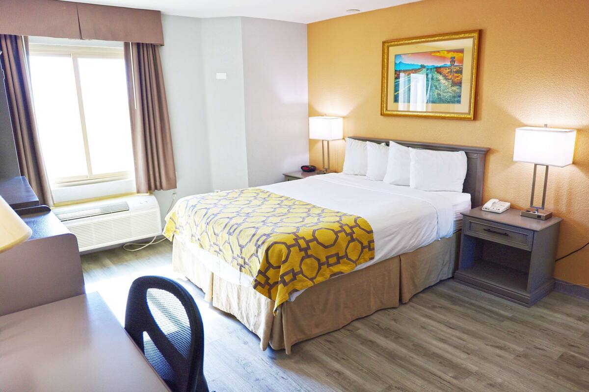 A hotel room with a bed, two bedside lamps, a desk with a chair, a window with curtains, and a painting on the wall.
