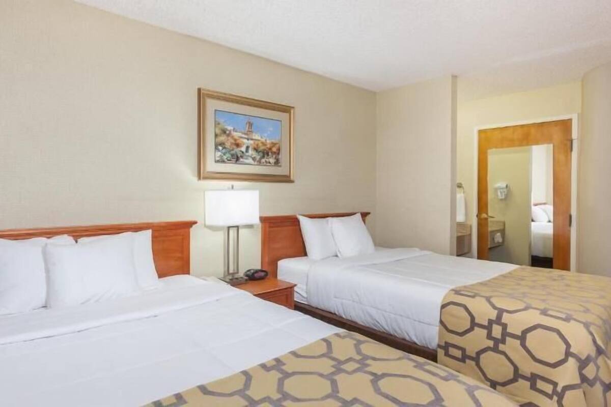 A hotel room with two beds, a painting on the wall, a bedside table with a lamp, and a door leading to a bathroom.