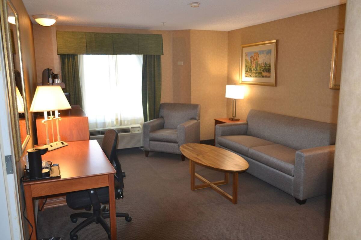 A hotel room with a desk, two armchairs, a sofa, a coffee table, a lamp, a painting, and a window with curtains.