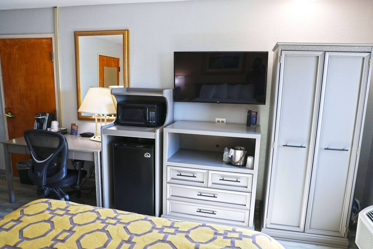 A hotel room with a bed, a desk with a computer, a microwave, a television, a wardrobe, and a chair.