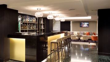 A modern bar area with a black countertop, bar stools, and a shelf with bottles. There is a seating area with a sofa and chairs in the background.