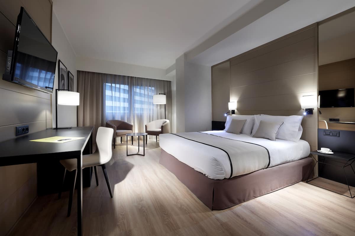Ein modernes Hotelzimmer mit einem großen Bett, einem Schreibtisch, einem Sessel, einem Fernseher und einem Nachttisch mit Lampe.