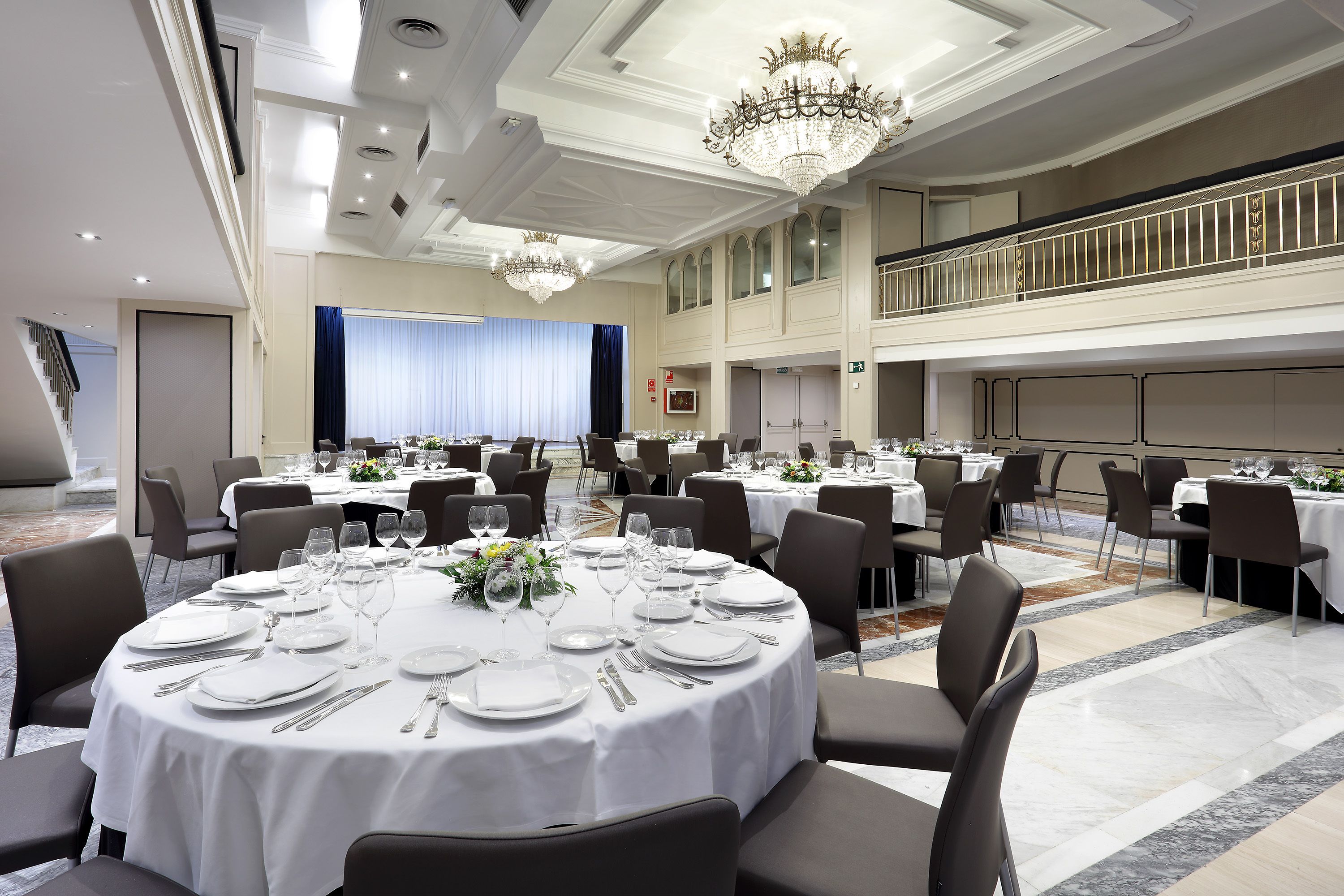 A banquet hall with round tables set for dining, a balcony with a railing, and a chandelier.