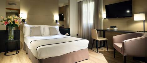 Una habitación de hotel con cama, mesitas de noche, escritorio, silla, televisor y un espejo grande.