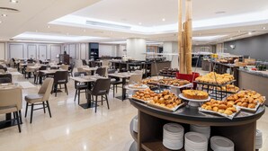 Un espace repas moderne dans un hôtel, avec de nombreuses tables et chaises, un buffet proposant diverses pâtisseries et desserts, et une grande colonne au centre.