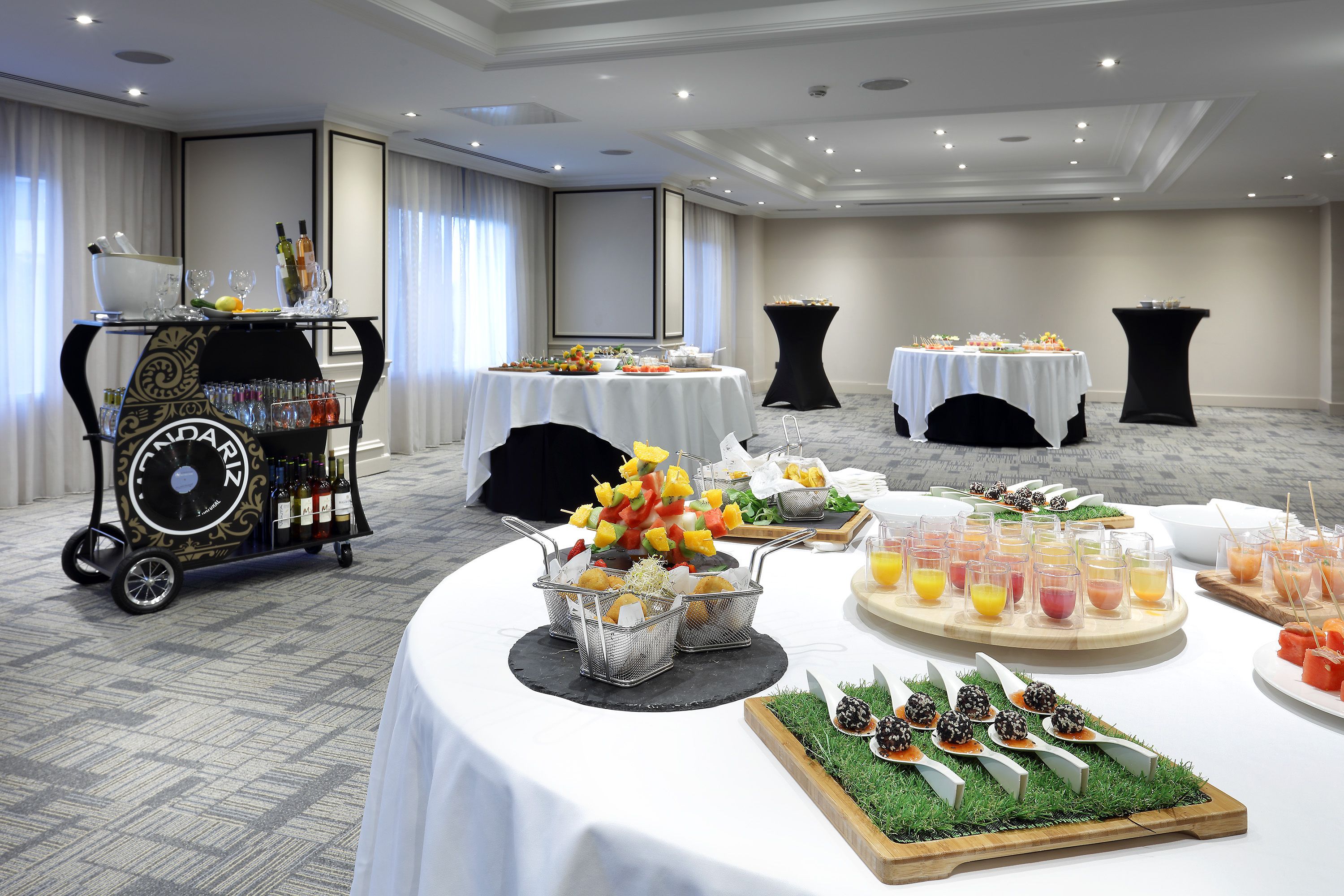 Disposición de un bufé con diversas opciones de comida y bebida, incluyendo brochetas de fruta, ensaladas y refrescos, dispuestas sobre mesas con manteles blancos en una sala amplia.