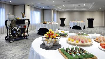 Disposición de un bufé con diversas opciones de comida y bebida, incluyendo brochetas de fruta, ensaladas y refrescos, dispuestas sobre mesas con manteles blancos en una sala amplia.