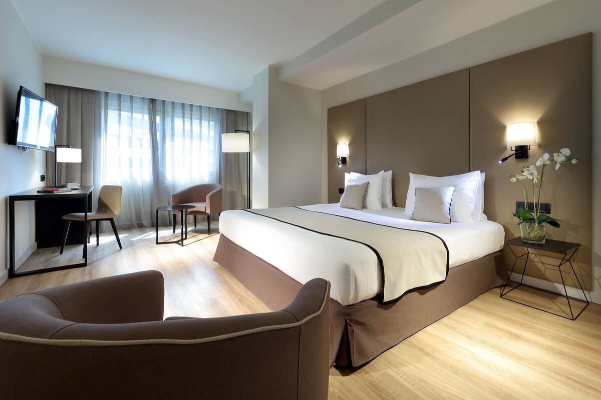 Ein Hotelzimmer mit einem großen Bett, einem Schreibtisch mit Stuhl, einem Fernseher und einem kleinen Nachttisch mit Lampe und Orchidee.