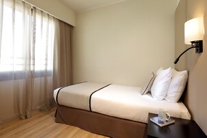 A hotel room with a bed, a bedside table with a lamp, and a window with sheer curtains.