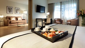 Une chambre d’hôtel comprenant un lit, une table basse avec des articles pour le petit-déjeuner, une télévision et un coin salon avec des fauteuils.