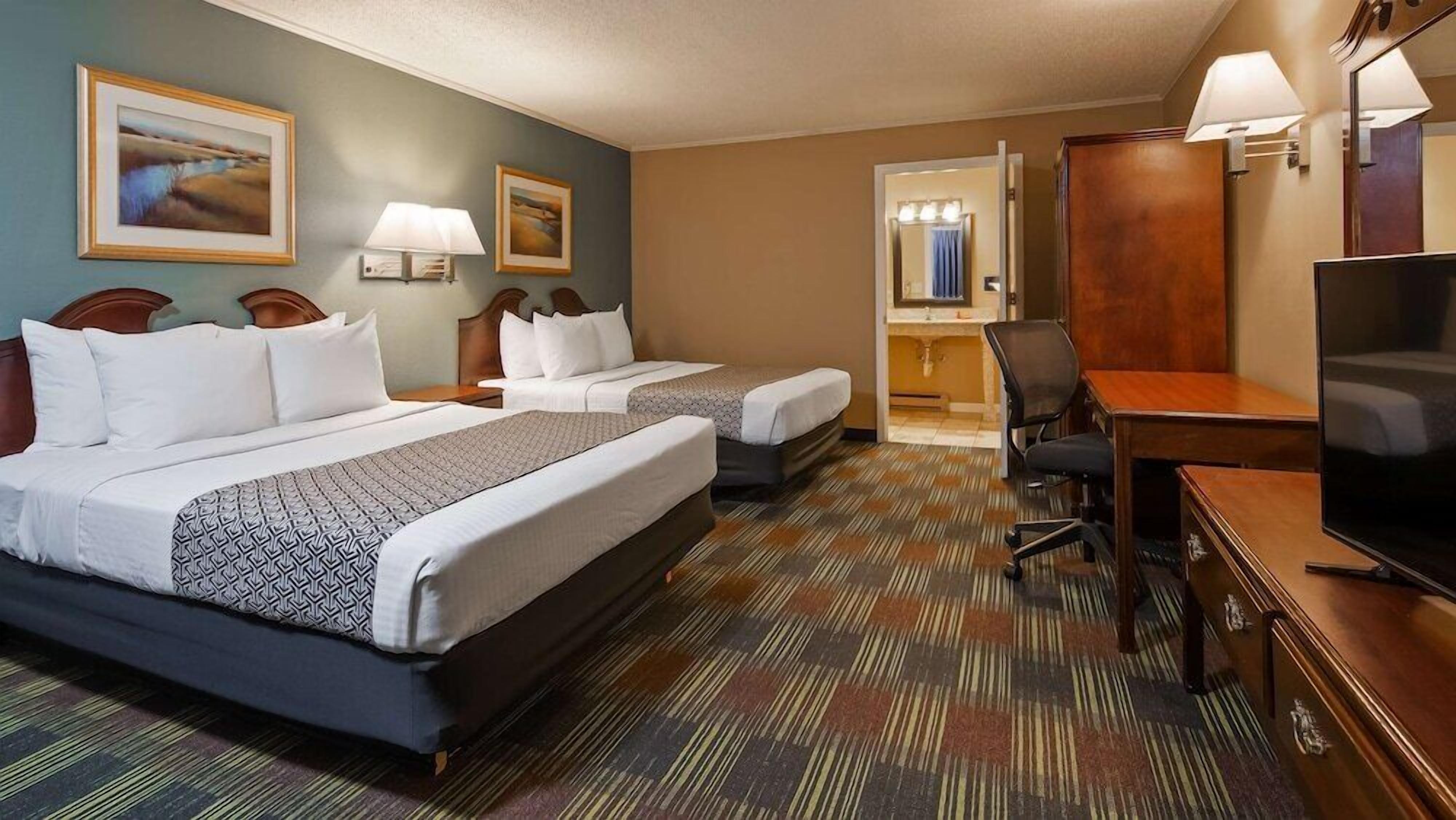 A hotel room with two beds, a desk, a chair, a TV, and a bathroom visible through an open door.