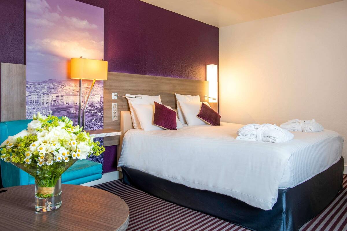 A hotel room with a bed, a nightstand, a lamp, a vase with flowers, and a cityscape painting on the wall.