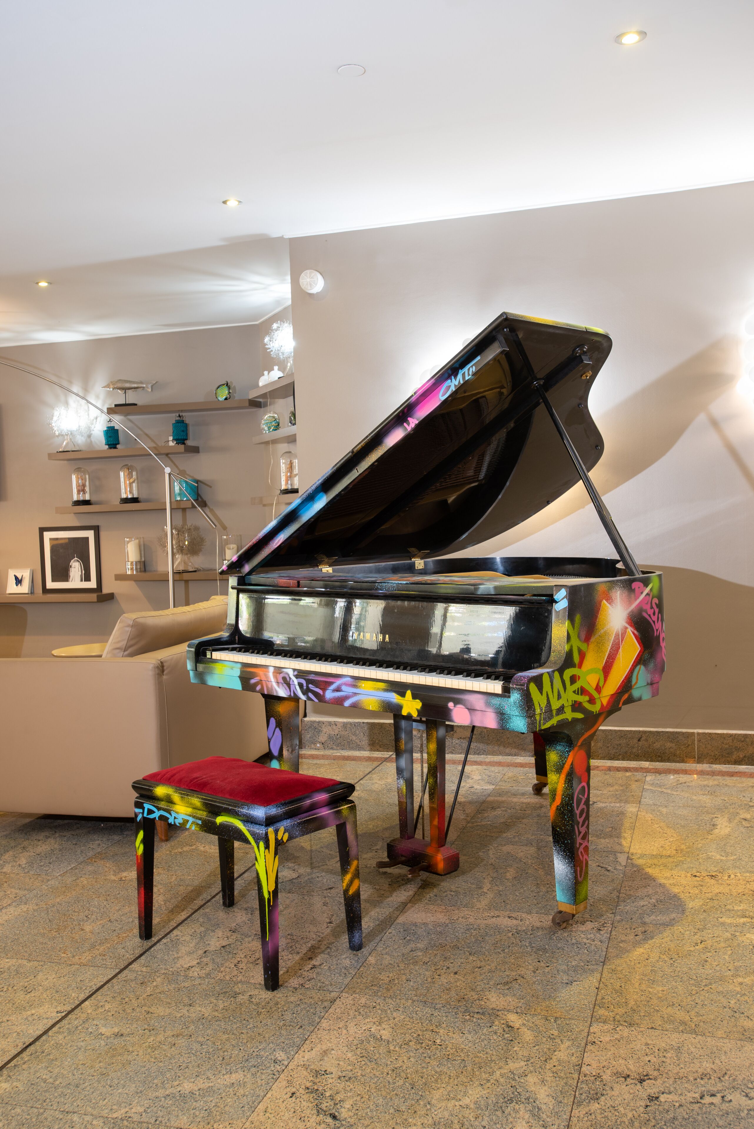 Un piano de cola con un diseño colorido en una habitación bien iluminada, con un sofá y estanterías decorativas.